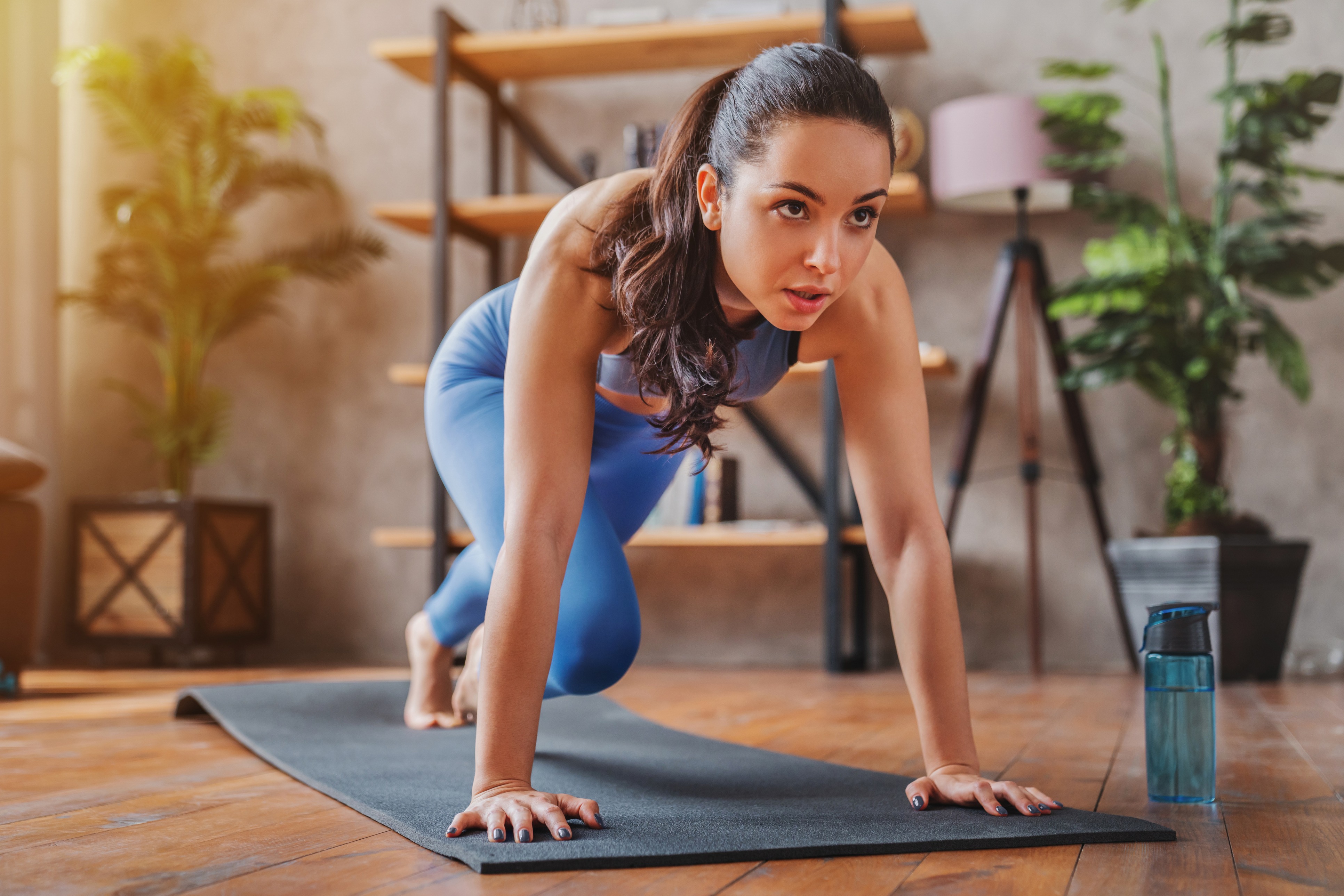Woman in a focused workout pose on a yoga mat at home with a water bottle nearby