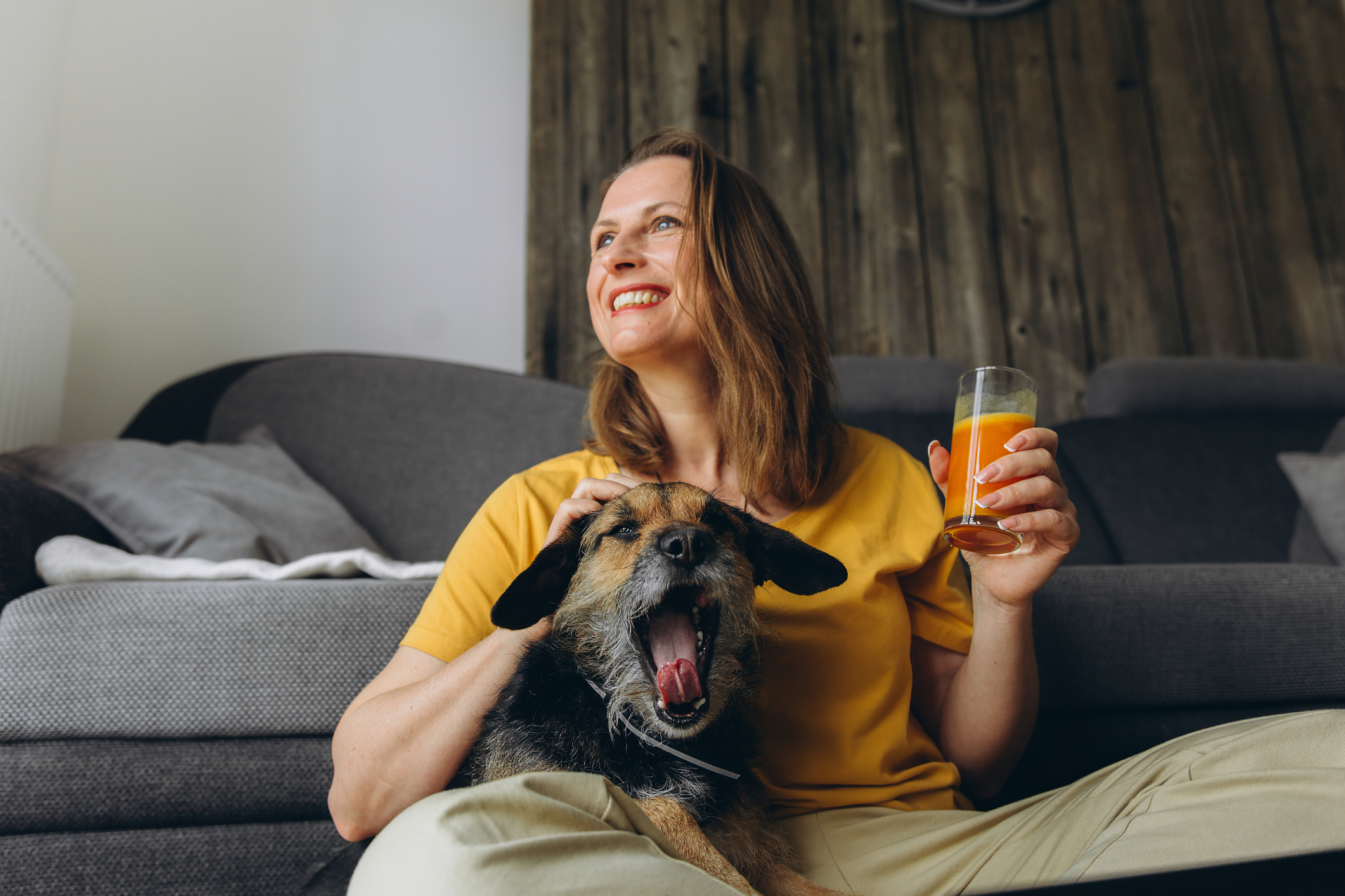 Smiling woman in a yellow shirt relaxing at home with her dog and a glass of fresh juice, staying hydrated and well during cold season